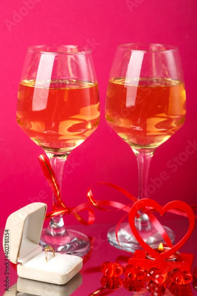 Fototapeta Two glasses of champagne tied with red ribbons on a pink background next to a ring in a box and a candle in a candlestick.