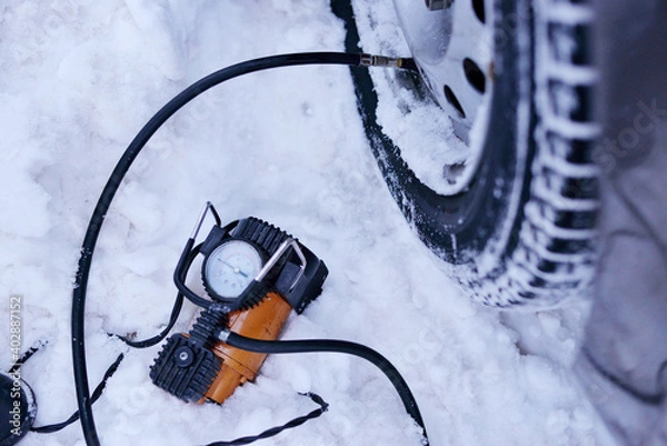 Obraz car pump compressor pumps up a flat tire of a car in winter on a snowy path in the forest close-up. Broken cars concept.