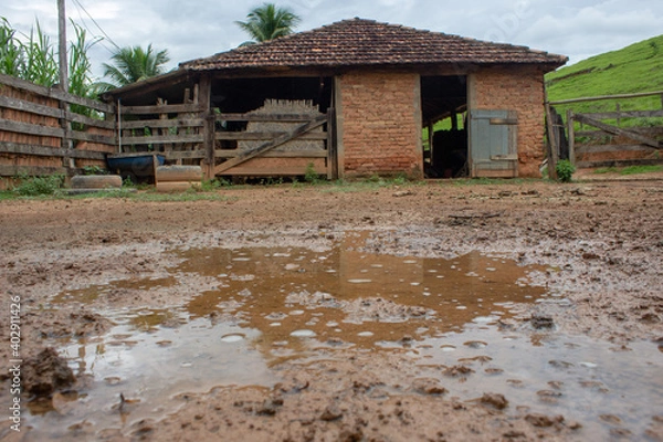 Fototapeta A kind of barn on a corral, after a heavy rain with the ground full of puddles. In the background there are a green pasture and blue sky