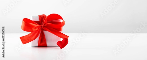 Fototapeta Gift box with red bow ribbon and red paper heart on white table for Valentines day with copyspace.