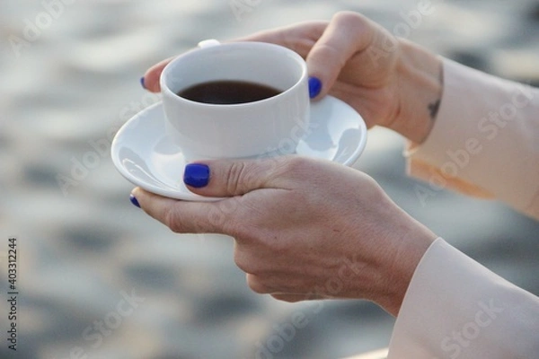 Obraz a white cup of coffee with a saucer in the graceful hands of a woman with a classic blue manicure and in beige clothes