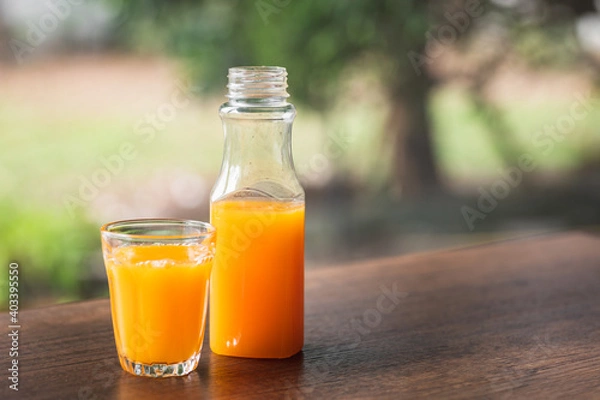 Obraz Fresh orange juice in no label clear plastic bottle in small glass on wooden table, natural trees blur in the background. With copy space.