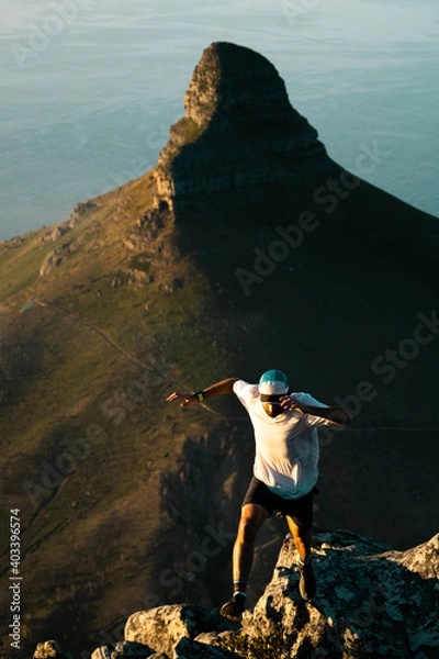 Fototapeta A young fit trail runner jumping across a steep ridge high in the mountains above Cape Town at sunset