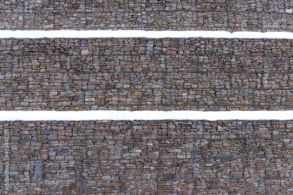 Fototapeta Metal Cage filled with rocks and snow texture . Stone wall with metal grid as background.  Gabion net