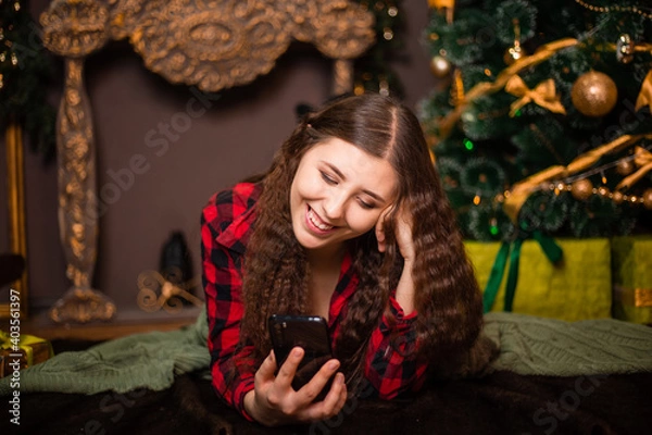 Fototapeta An attractive woman lies on a fur carpet with a smartphone in her hands against the background of a Christmas tree with gifts and a fireplace.