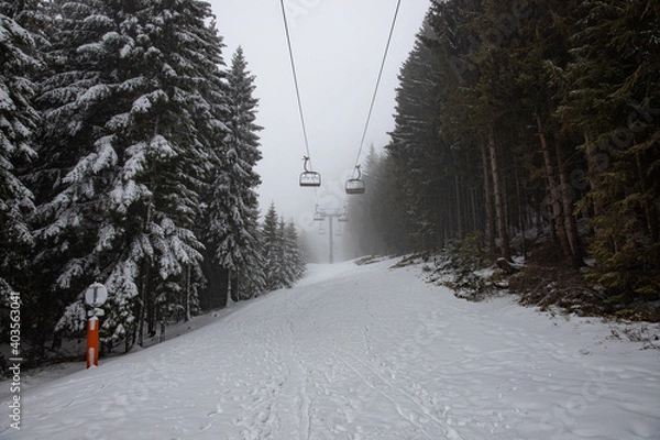 Obraz Empty Ski Lift on a snowy winter landscape, with snow covered mountains and big pine and fir trees, in the fog. The chairlift has no people on it.