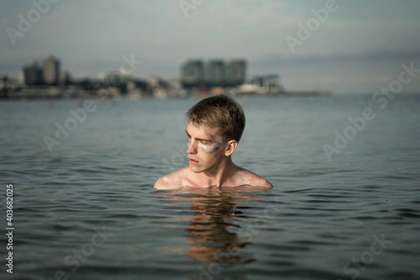 Obraz portrait of a non-binary young man with sparkles on his face, standing in the sea, in the background you can see the city