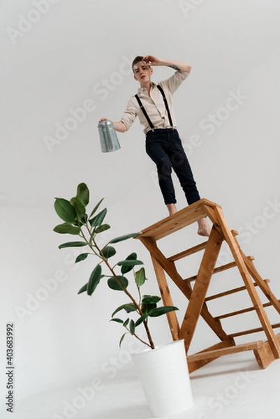 Fototapeta a guy stands on a wooden decorative ladder in a shirt watering a growing tree in a white pot from a garden watering can, an angle from the bottom
