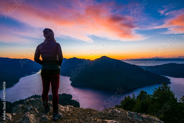 Fototapeta Adventurous athletic woman hiker standing on a mountain top watching the sunset over mountains and a beautiful lake.