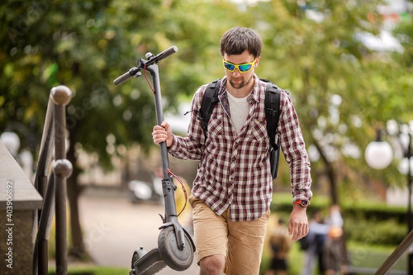 Fototapeta Modern city dweller caucasian man carries electric scooter over obstacle in park while driving around the city. Theme of convenient functional public space, city infrastructure and inclusiveness