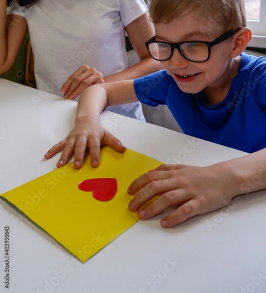 Fototapeta Mother and happy son happy make a Mother's Day card sitting together at a table with paper and a red heart. A happy child with glasses made a card with his own hands for Valentine's Day, Father's Day.