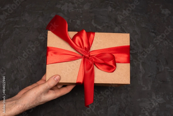 Fototapeta A gift box with a red bow is held in the hands of a man on a dark background