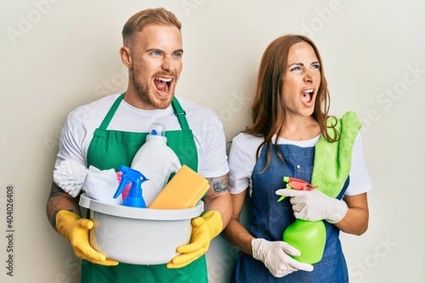 Fototapeta Young couple of girlfriend and boyfriend wearing apron holding products and cleaning spray angry and mad screaming frustrated and furious, shouting with anger. rage and aggressive concept.