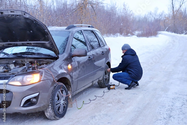 Obraz Man checking air pressure and filling air in the tires with Car eletric pump on road in winter on a snowy path in the forest using a pump. Broken cars concept