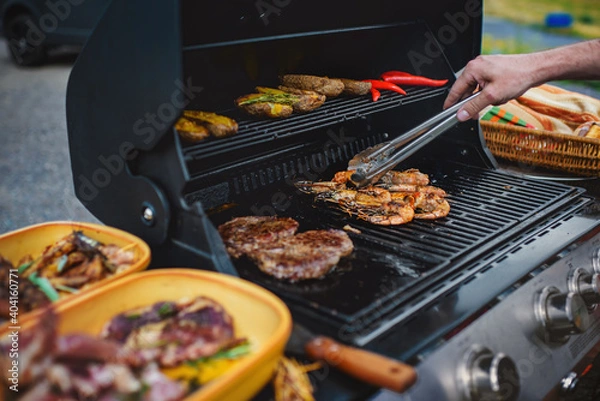 Fototapeta Male hand with tongs turning the giant tiger prawn on an outdoor grill, where other types of meat and vegetables are also grilled.