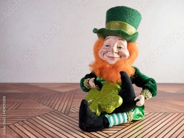 Obraz St Patrick’s Day leprechaun with a green outfit and green hat sitting on a wooden surface with a reddish orange fluffy beard, holding a glittery green four leaf clover.
