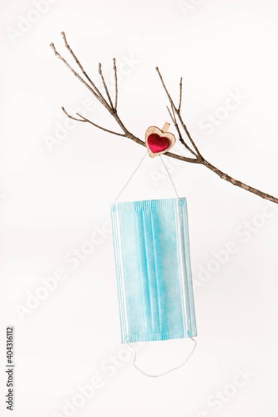 Obraz Medical social blue mask against COVID19 coronavirus 2021 hangs on a bare branch of a tree for Valentine's Day pinned down with a red heart shaped clothespin. White background isolated