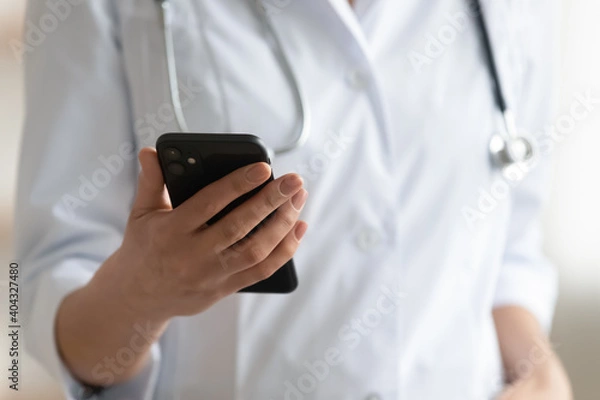 Fototapeta Close up young nurse or doctor holding cellphone in hands, reading message or web surfing information online. Female physician general practitioner using mobile applications, telemedicine concept.