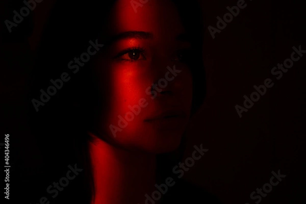 Fototapeta The close view of young woman's face illuminated with red light in the dark. Depressed woman suffering because of isolation during coronavirus pandemic, she stays in home. She is sad and lonely.