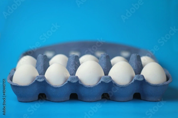 Fototapeta White chicken eggs in a color paper tray on a blue surface, selective focus