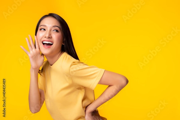 Fototapeta Surprised happy beauty asian woman looking copy space in excitement Expressive facial expressions Presenting some product. Beautiful girl act like a telling secret Isolated on yellow background
