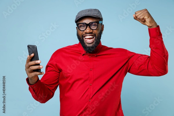 Fototapeta Joyful young bearded african american man 20s in casual red shirt eyeglasses cap doing winner gesture using mobile cell phone typing sms message isolated on pastel blue background studio portrait.