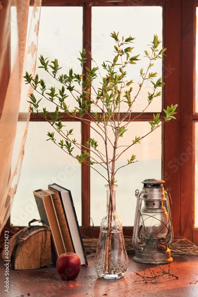 Fototapeta spring twig in a transparent glass vase with a kerosene lamp and books on the background of the window