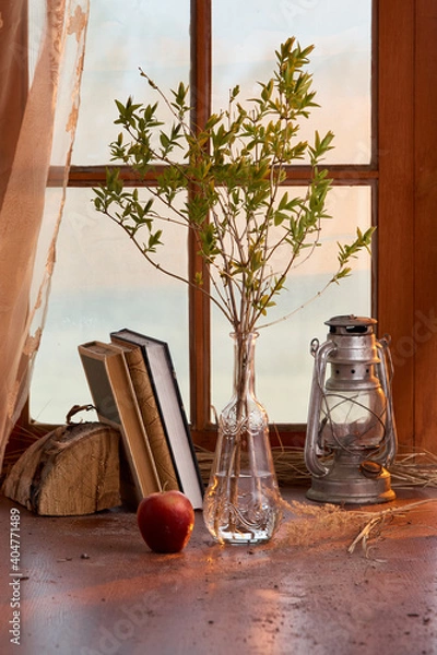 Fototapeta spring twig in a transparent glass vase with a kerosene lamp and books on the background of the window