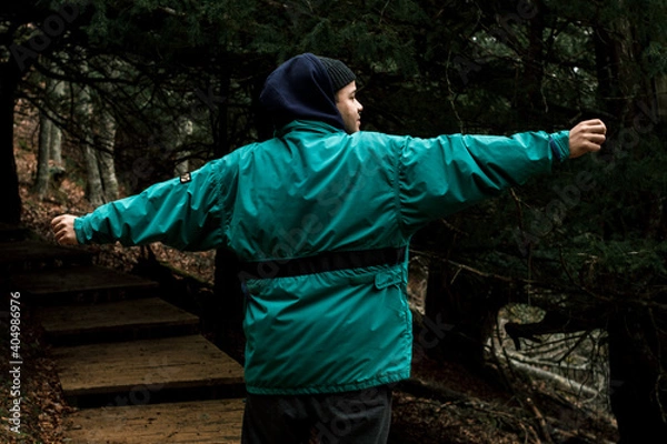 Fototapeta The young hiker with blue coat and hood extends his arms while walking along a wooden path among the trees