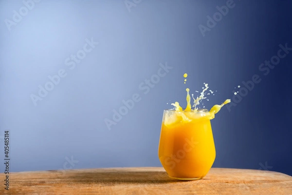 Fototapeta Glass of fresh orange juice splash on on wooden table and blue background. Dynamic pour of fruit juice in the morning. Space for text