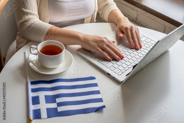 Obraz Lonely woman freelancer with flag of Greece enjoying having breakfast with cup of coffee working on laptop sitting near window in cafe at morning.