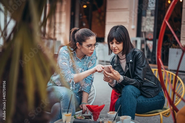 Fototapeta Two beautiful women in cafe outdoors looking at something on mobile phone. Two female using mobile phone application shopping online and enjoying coffee outside in city
