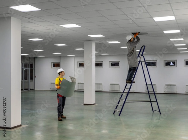 Fototapeta Workers on a ladder check the electrical wires on the ceiling. Workers in overalls check the correctness of the wiring under the ceiling in the office space. Check with the plan.