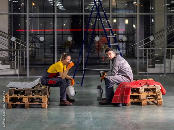 Fototapeta Workers' lunch break. Workers in overalls rest and drink tea while sitting on wooden pallets in the building. Rest in the middle of the working day.