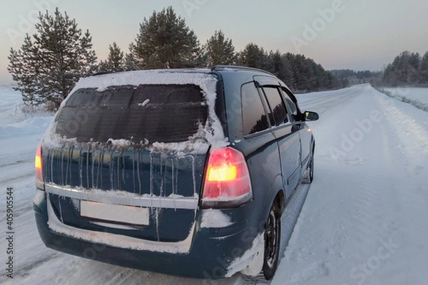 Fototapeta a frozen car stands on the side of a slippery winter road at dusk. Orange warning lights are on. The rear window was covered with ice, snow and icicles. Dangerous long journeys in the cold.