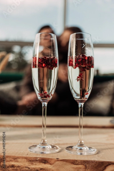 Fototapeta Happy Valentines day celebration concept. Close up of 2 two glasses with sparkling champagne and red heart shaped confetti. Love potion in glass. Loving couple blurred on background