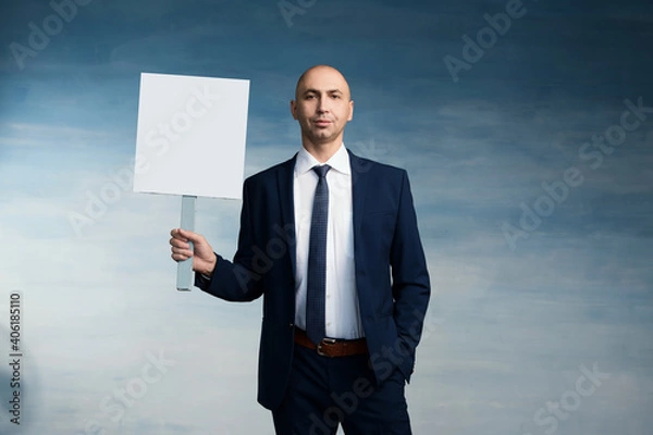 Fototapeta A bald man in a business blue suit spectacle and tie stands with holding a poster in his hand on a blue background