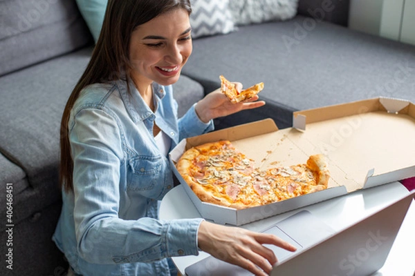 Fototapeta A young smiling woman is working on her laptop and eating pizza at home.