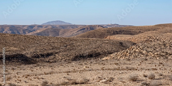 Fototapeta a rugged barren mountainous desert landscape west of the makhtesh ramon crater with an israeli border outpost and camel hump mountain in the sinai in the background against a clear blue sky