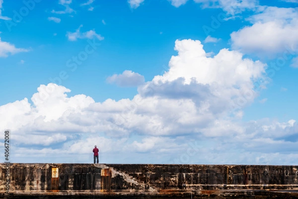 Fototapeta A single man stands on the harbour walls looking outwards towards the open sea and the vast expanse of a lovely sky.