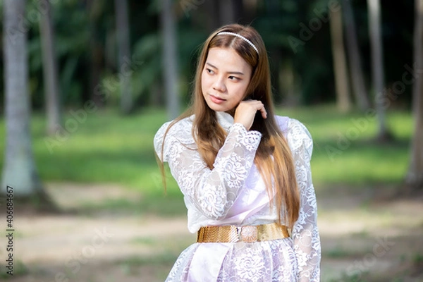 Fototapeta Asian woman in a Thai dress is standing in the garden with green tree background. Beautiful Thai girl looking something.