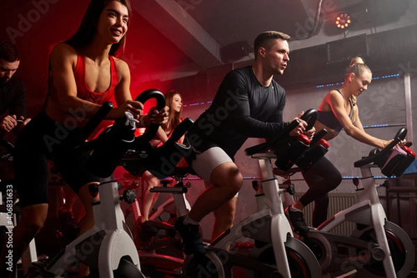 Fototapeta people biking in spinning class at modern gym, exercising on stationary bike. group of caucasian people athletes training on exercise bike