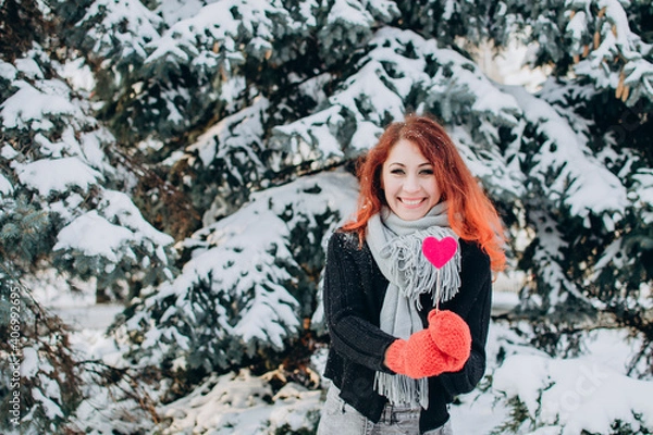 Fototapeta A girl with red hair holds a heart on a stick in her hands against a background of trees and snow. Valentine's day, winter, love, feelings, discounts and promotions.