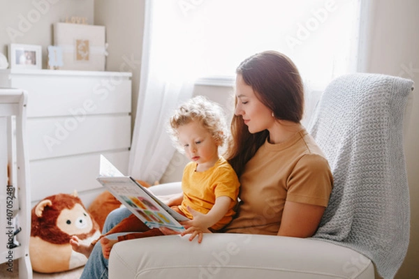Fototapeta Mom reading book with baby boy toddler at home. Early age children education, development. Mother and child kid spending time together. Family authentic candid lifestyle.