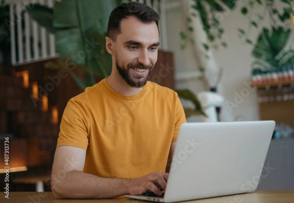 Fototapeta Attractive smiling man using laptop computer, working from home. Portrait of young freelancer copywriter typing, sitting at workplace. Student studying, distance learning, searching, online education