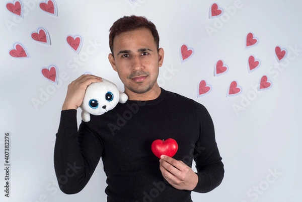 Fototapeta Young man holding a red love heart on his chest and a baby seal plush on his shoulder, with red paper hearts flying behind him. Concept of love, valentine 