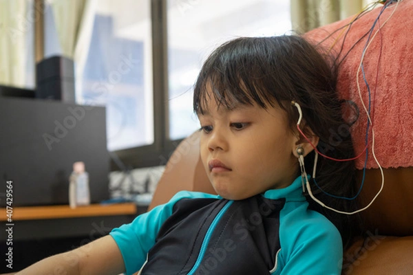 Fototapeta A little girl is wearing a hearing aid and is doing a neurofeedback exam. Making EEG electrodes.