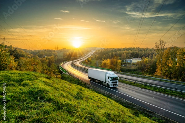 Fototapeta White truck driving on the highway winding through forested landscape in autumn colors at sunset