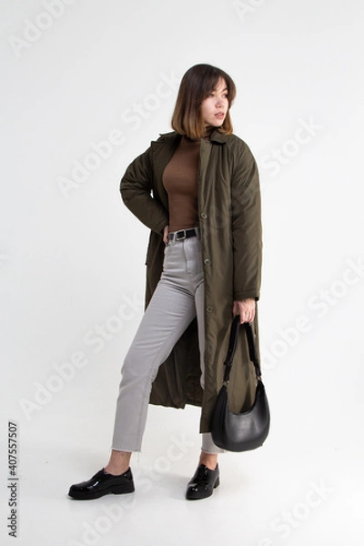 Obraz Young attractive woman in long green coat with black handbag on white background. Model in gray short pants, brown sweater and black shoes looking to the right
