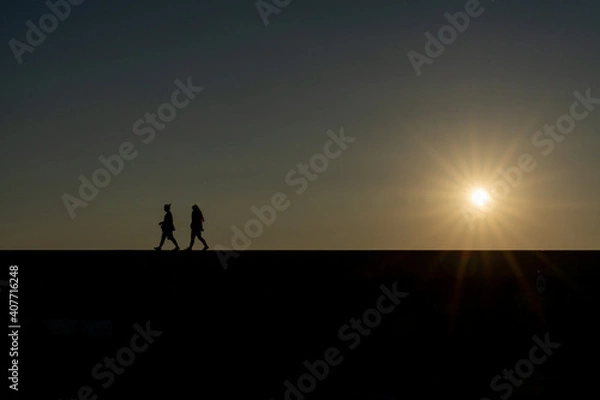 Fototapeta silhouette of two women walking on a wall with the setting sun and evening sky